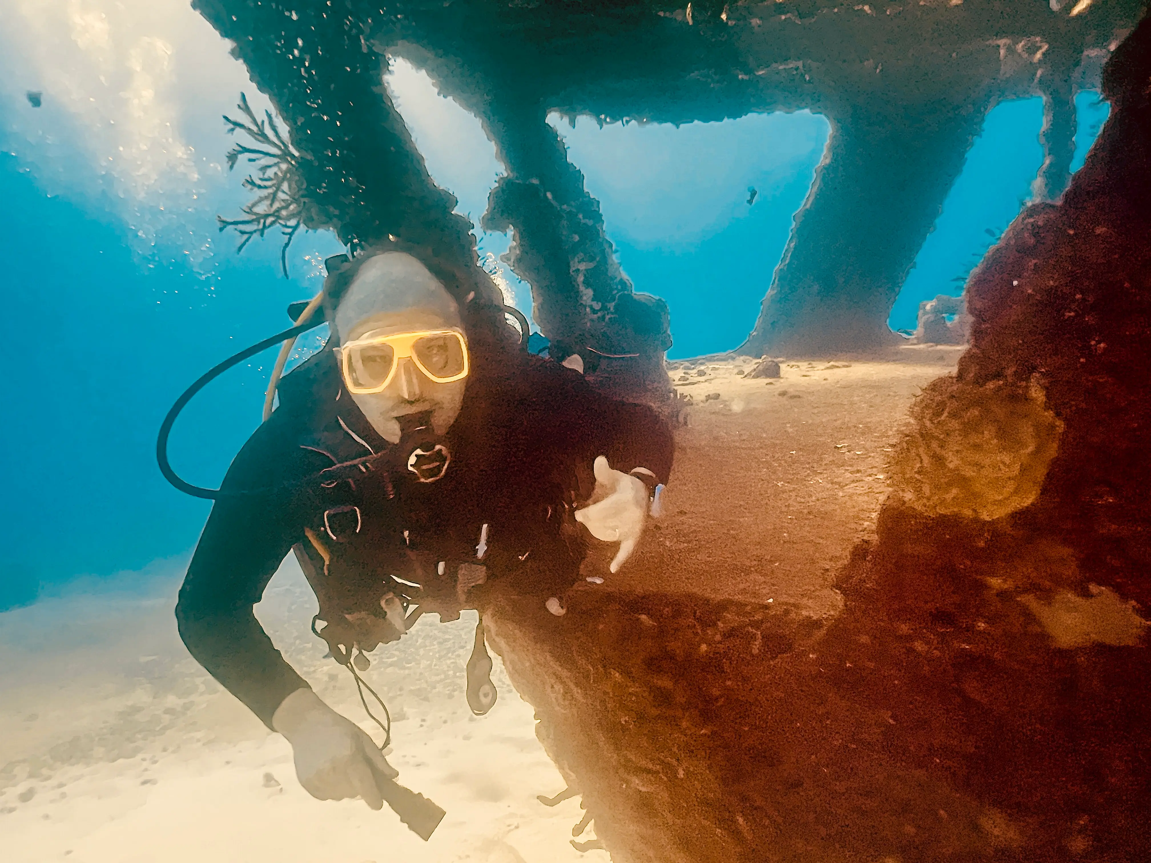 A diver with a lamp explores a wreck, with bubbles and light filtering through the structure in clear water.